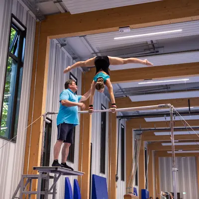 Ein Trainer hilft einer Turnerin der Turngemeinde Landshut beim Handstand am Stufenbarren in einer Sporthalle.