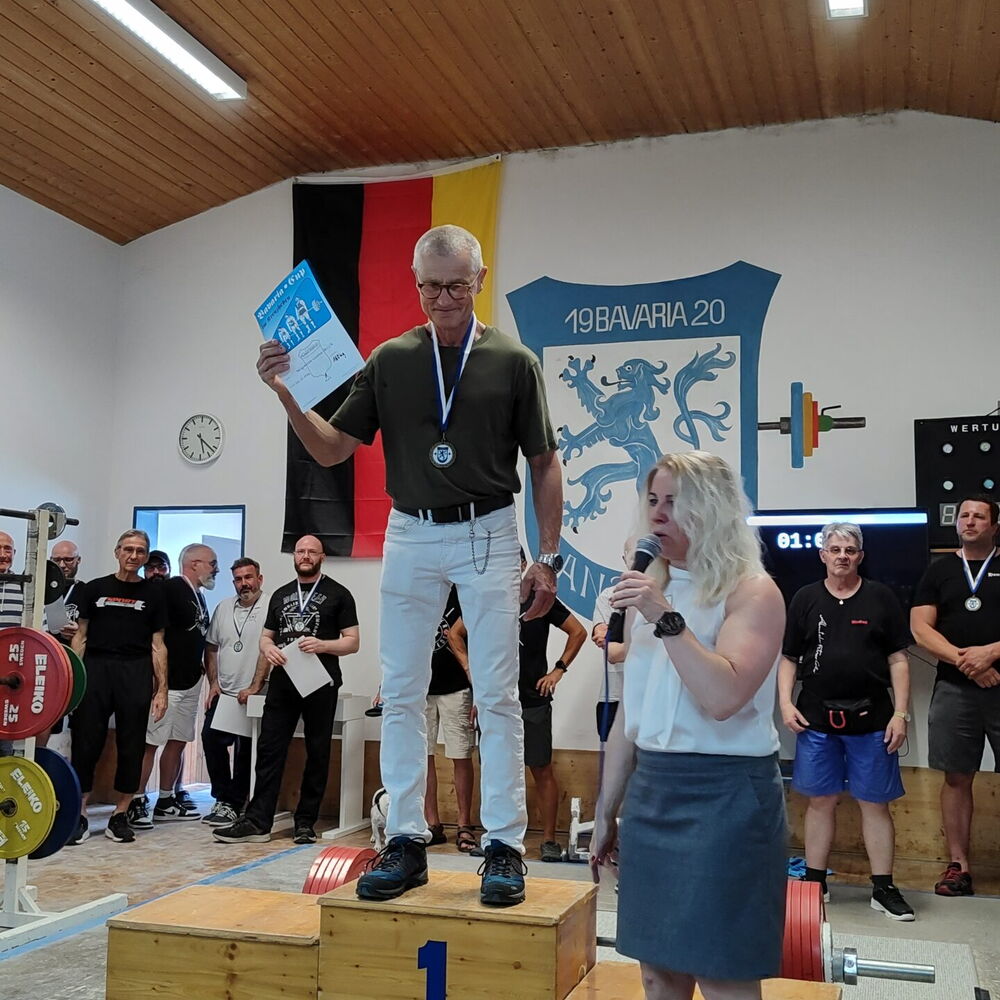 Ein Mann steht mit Medaille auf dem Siegerpodest bei einem Turngemeinde-Wettbewerb in Landshut, während eine Frau spricht.