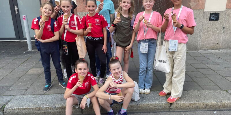 Die kleine TeamGym-Mannschaft der Turngemeinde genießt fröhlich Eis in der Sonne vor einem Gebäude in Landshut.