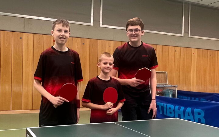 Drei junge Tischtennisspieler der Turngemeinde Landshut posieren stolz mit Schlägern in der Sporthalle des Vereins.