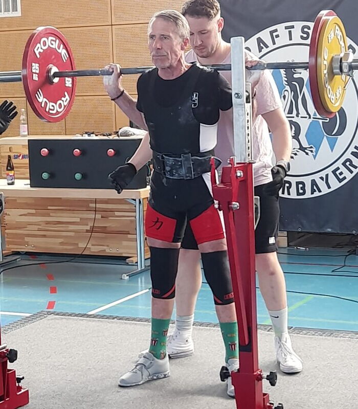 Ein Sportler der Turngemeinde hebt eine Langhantel bei einem Wettkampf in der Halle, unterstützt von einem Spotter.