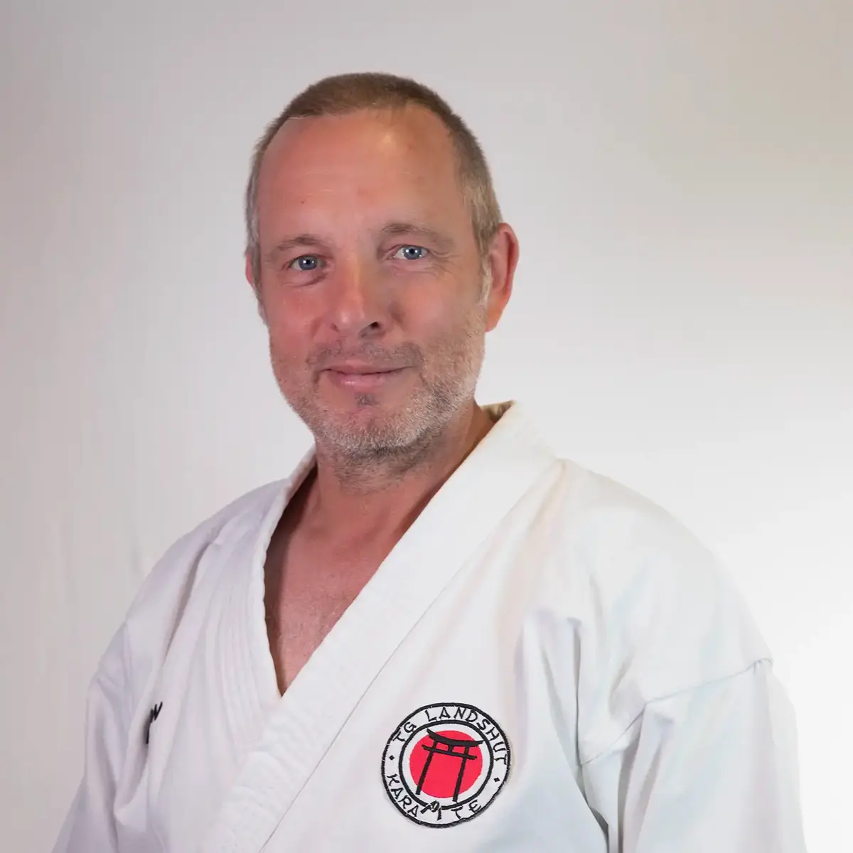 Ein Mann im Karateanzug mit TGL-Logo steht stolz vor weißem Hintergrund, repräsentiert den Sportverein Landshut.