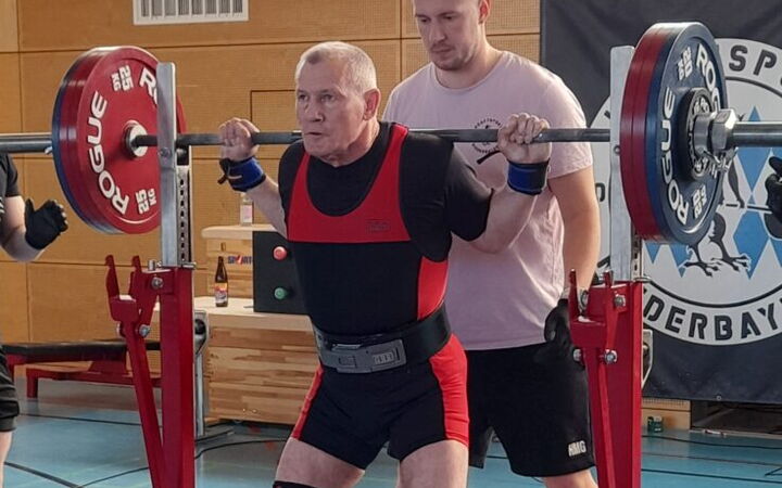 Ein Sportler der Turngemeinde hebt Gewichte, unterstützt von einem Trainer in einer Sporthalle.