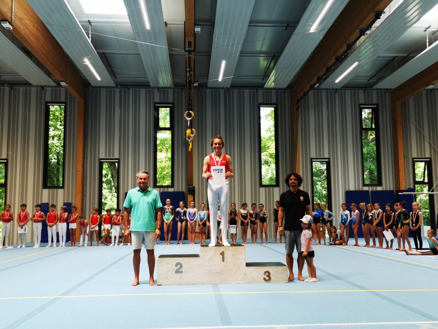Ein Junge steht stolz auf dem Siegerpodest in der Turnhalle des Turnvereins Landshut, umgeben von anderen jungen Sportlern.