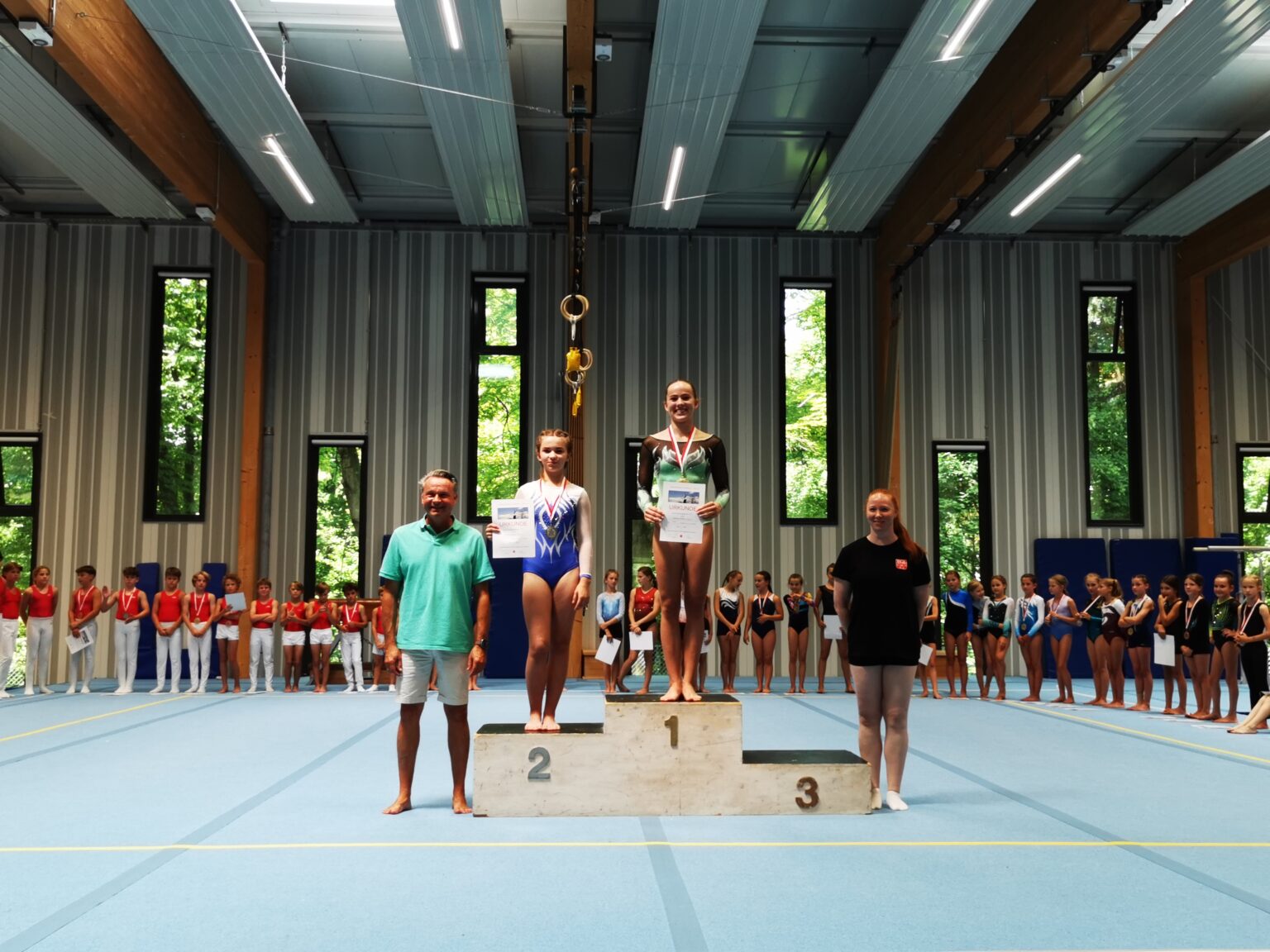 Zwei Mädchen stehen auf einem Podest in einer Turnhalle, umgeben von anderen Teilnehmern des Turnvereins Landshut.