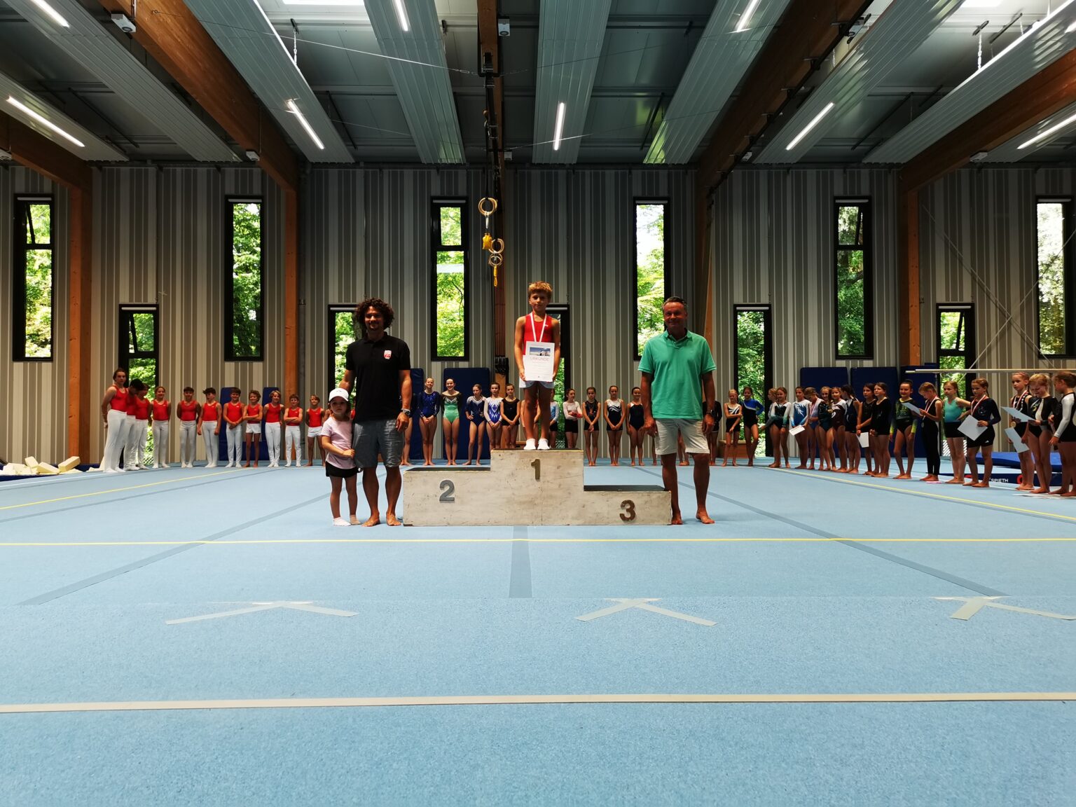Ein Junge steht auf dem Siegerpodest in einer Turnhalle, umgeben von anderen Sportlern des Vereins Turngemeinde Landshut.